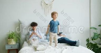 快乐的孩子在床上蹦蹦跳跳，而妈妈在用智能手机，爸爸拿着平板电脑